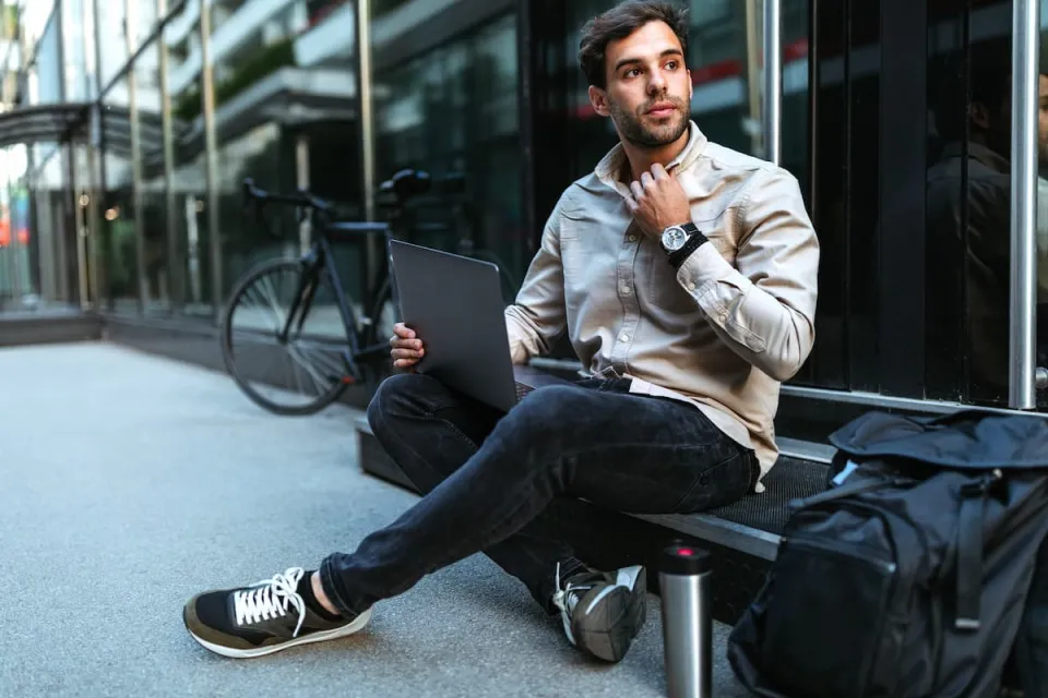jonge man zit buiten op een bankje met laptop op schoot en kijkt in de verte